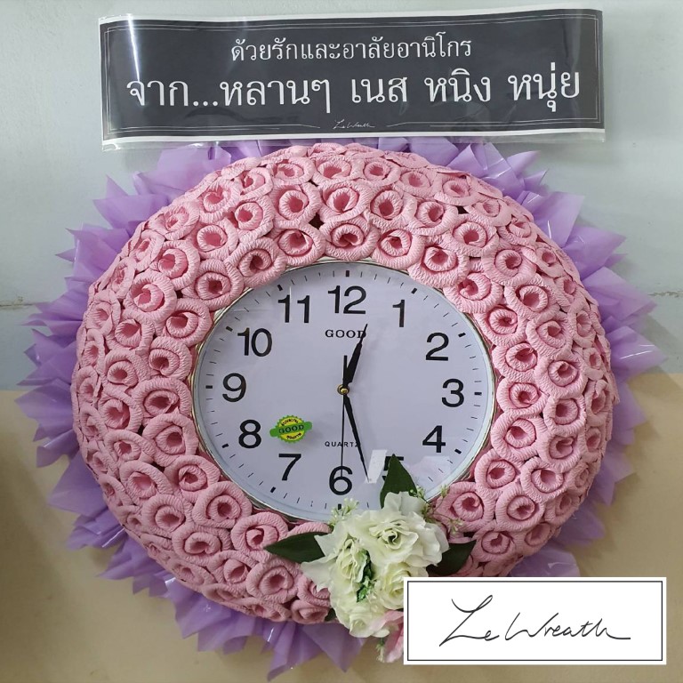พวงหรีดนาฬิกาตกแต่งด้วยดอกไม้จันทน์โทนสีชมพู ตัดกับพิ้นหลังสีม่วงอย่างสวยงาม