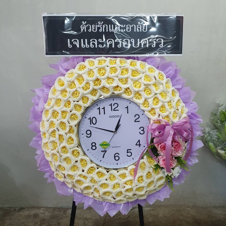พวงหรีดดอกไม้จันทน์นาฬิกา โทนสีสดใส นำนาฬิกาขนาดเส้นผ่าศูนย์กลางประมาณ 60-80 ซม. มาตกแต่งดอกไม้จันทน์สีเหลือง ตัดกับพื้นหลังสีฟ้า
